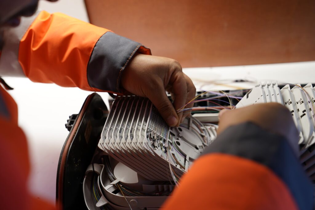 Mains de techniciens ENSIO en veste orange manipulant les fibres optiques dans un boîtier de raccordement aérien ouvert