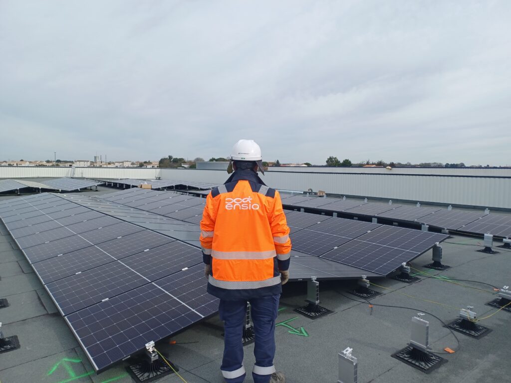 Technicien ENSIO de dos en veste orange sur une toiture plate inspectant une installation de panneaux solaires photovoltaïques