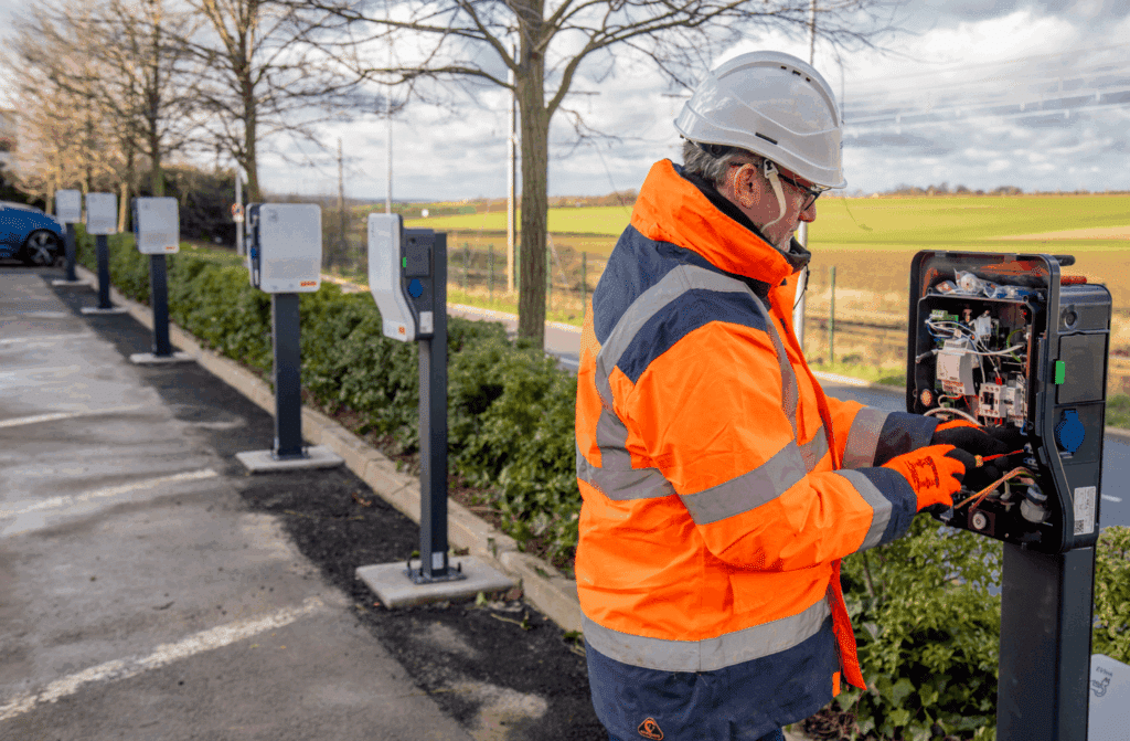 Technicien en veste orange et casque blanc raccordant une borne de recharge pour véhicules électriques, avec une rangée de bornes IRVE installées en arrière-plan