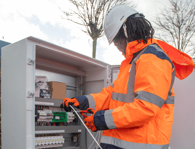 Technicien en veste orange et casque blanc raccordant un coffret électrique lors d'une installation de borne de recharge pour véhicules électriques