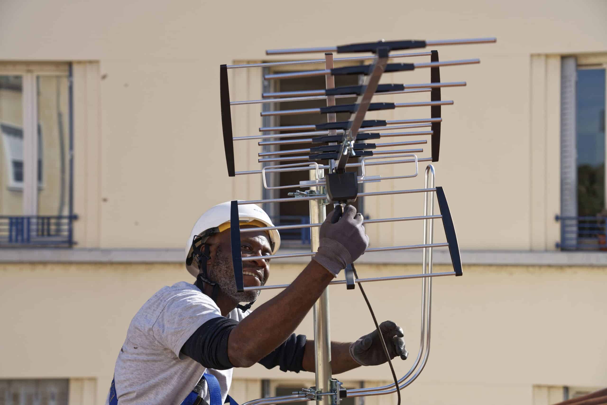 Technicien souriant en casque blanc installant une antenne râteau sur un mât en milieu urbain, en façade de bâtiment résidentiel