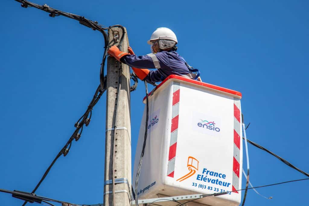 Technicien ENSIO en nacelle élévatrice intervenant en hauteur sur un poteau électrique pour un branchement au réseau, sur fond de ciel bleu
