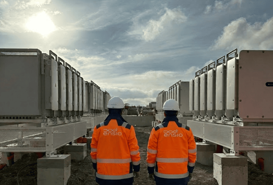 Deux techniciens ENSIO de dos en veste orange et casque blanc face à une allée de containers de stockage d'énergie sur un site industriel