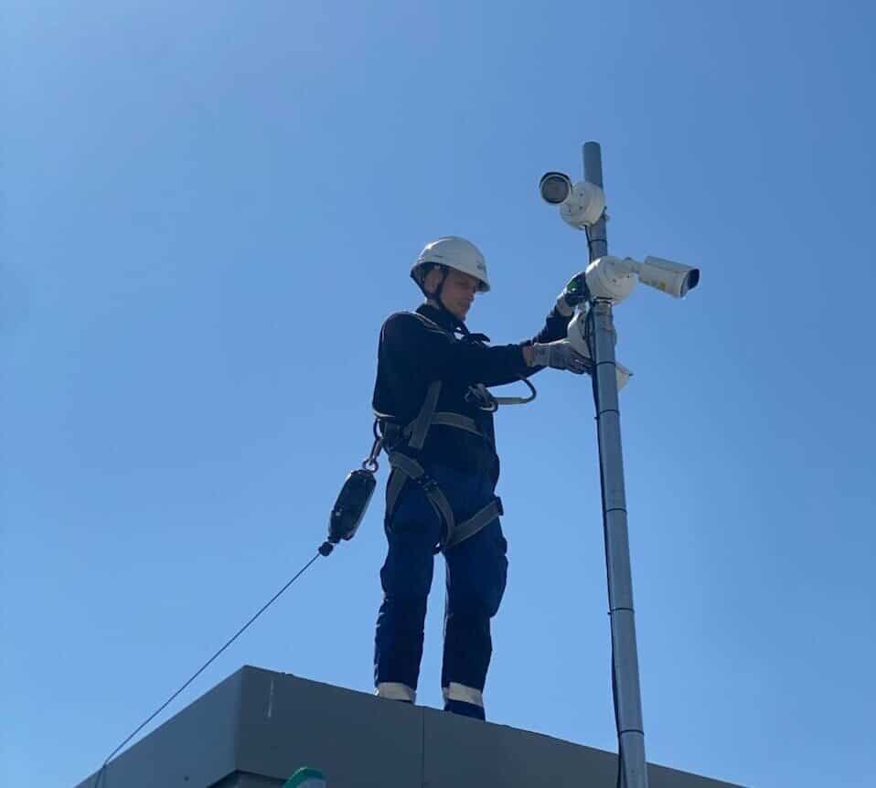 Technicien équipé d'un harnais de sécurité installant des caméras de vidéosurveillance sur un mât en toiture, sur fond de ciel bleu