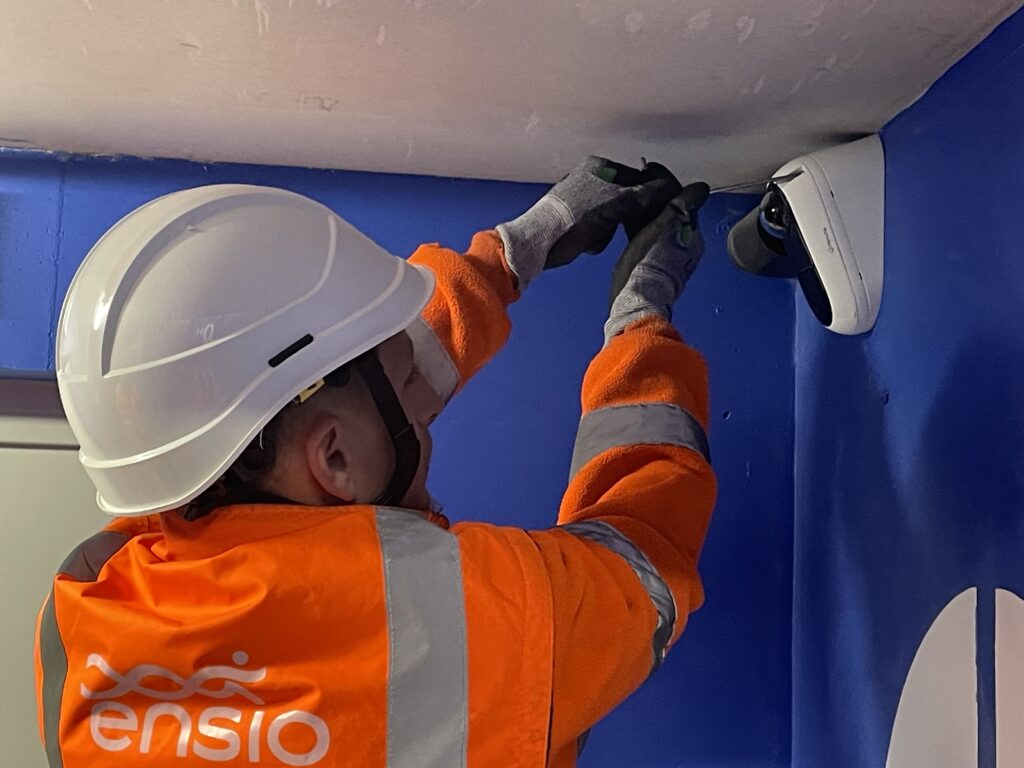 Technicienne ENSIO en veste orange et casque blanc fixant une caméra de vidéosurveillance dome au plafond en intérieur
